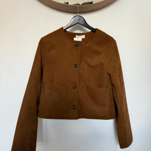 Mockajacka- H&M, S (ny med prislapp) - Snygg brun mockajacka från H&M med rak, croppad passform och stora fickor framtill. Jackan har rund halsringning, knäppning med svarta knappar och långa ärmar. Perfekt för dig som gillar stilren och enkel design.