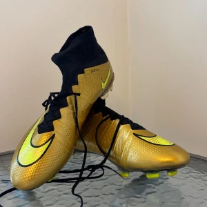 Guldiga Nike fotbollsskor med högt skaft - Säljer ett par riktigt snygga fotbollsskor från Nike i metallic guld med svarta detaljer och högt strumpliknande skaft. Skorna har svarta snören, tydliga neongula Nike-loggor och genomskinlig sula med gula dobbar. Perfekta för dig som vill sticka ut på planen. 