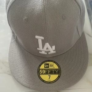 Grå LA Dodgers keps från New Era - Snygg grå 59FIFTY keps från New Era med LA Dodgers broderat i vitt framtill. Klassisk flat peak och broderade ventilationshål. Kepsen har flera broderade loggor i vitt på sidorna och bak. Perfekt för dig som gillar streetwear och sportig stil.
