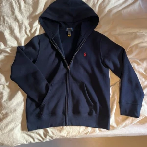 Marinblå hoodie från Polo Ralph Lauren - Snygg marinblå hoodie från Polo Ralph Lauren med dragkedja och huva. Klassisk röd broderad logga på bröstet och två stora fickor framtill. Perfekt för chill dagar och enkel att matcha med jeans eller joggers har används endast 2 gånger .