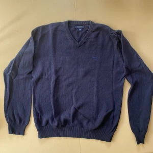 Mörkblå stickad tröja från GANT - Klassisk mörkblå stickad tröja från GANT med v-ringning och diskret broderad logga på bröstet. Tröjan har långa ärmar och ribbade muddar vid ärmslut och nederkant. Perfekt för dig som gillar stilren och tidlös design.