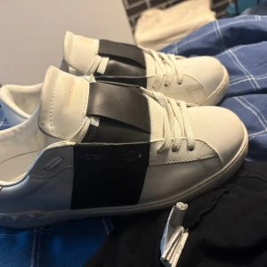 Valentino Garavani Open sneakers vit/svart - Säljer ett par Valentino Garavani Open sneakers i vitt skinn med bred svart läderdetalj över mitten. Klassisk rund tå, vita snören och diskret Valentino-logga på plösen. Snygg och stilren design som sticker ut med den svarta kontrasten.