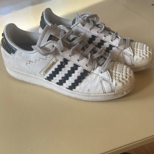 Adidas Superstar x LEGO sneakers vit/svart - Unika Adidas Superstar sneakers i samarbete med LEGO. Skorna är vita med svarta detaljer och har LEGO-inspirerade mönster på tå, häl och sidor. Klassisk snörning och platt sula. Tillverkade i skinn med coola zigzag-detaljer och tydlig LEGO branding.