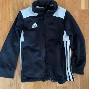 Svart Adidas vindjacka med vita detaljer - Sportig svart zip-tröja från Adidas. Jackan har dragkedja framtill, hög krage och Adidas-logga på bröstet. Perfekt för dig som gillar en clean och atletisk stil. STORLEK 122-128