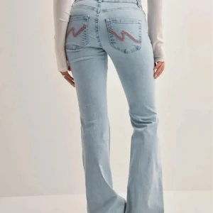 Ljusblå bootcut jeans med slitningar - Snygga ljusblå jeans med bootcut passform och slitna detaljer vid knäna. Jeansen har klassiska bakfickor med dekorativa sömmar i rött och normalhög midja. Perfekta för en avslappnad och trendig look.