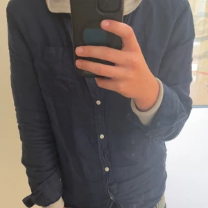 Mörkblå skjorta  - Snygg mörkblå skjorta med klassisk krage och vita knappar framtill. Skjortan har lång ärm och en enkel bröstficka. Perfekt att bära över en hoodie för en avslappnad stil. Materialet känns lätt och skönt mot huden.