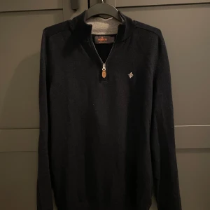 Mörkblå stickad tröja från Morris - Tvärfet mörkblå stickad tröja från Morris med halv dragkedja och liten broderad logga på bröstet. Sinnessjukt bra skick, iprincip helt nyskick, tröjan har hög krage och långa ärmar, perfekt för kyliga dagar. Klassisk och stilren design som funkar till det mesta.