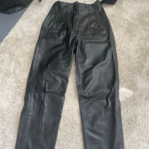 Svarta skinnbyxor från Zara XS - Snygga svarta skinnbyxor från Zara i storlek XS. Byxorna har hög midja, raka ben och stängs med dragkedja, knapp och hake. De har två fickor fram och är tillverkade i ett mjukt syntetmaterial med skinnlook.