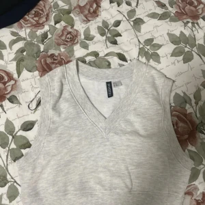 Ljusgrå v-ringad väst från H&M - Snygg ljusgrå väst från H&M i storlek XS. Västen har v-ringning och breda ribbade kanter vid hals och ärmhål. Perfekt att styla över en skjorta eller t-shirt för en trendig lager-på-lager-look.