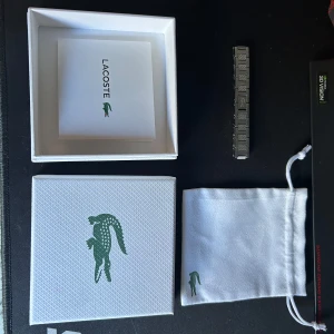 Lacoste silverfärgad armband  - Snygg och stilren klocka från Lacoste med silverfärgat armband i metall. Armbandet har en rektangulär länkdesign och är prytt med Lacostes ikoniska krokodillogo. Levereras med original ask och tygpåse med logga.