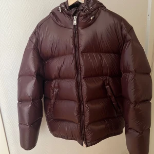 Brun pufferjacka med huva - Säljer en Zara  mörkbrun pufferjacka med huva och snörning. Jackan har dragkedja framtill, två sidofickor och en riktigt puffig look. Perfekt för kalla dagar och har en oversized passform. Materialet är glansigt syntet och jackan är långärmad.