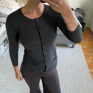 Mörkgrå kofta med knappar - Mörkgrå kofta med rund halsringning och knäppning framtill. Tröjan har två små fickor framtill och är tillverkad i ett mjukt, stickat material. Ärmarna är långa och passformen är figurnära, perfekt för lager-på-lager-stil.