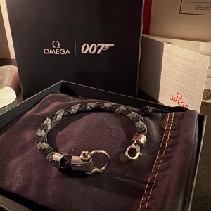 Omega 007 svart och grått armband - Exklusivt Omega 007 armband med flätad design i svart och grått. Snygga silverfärgade metalldetaljer och robust lås. Kommer i originalförpackning med Omega och 007-logga, perfekt för dig som gillar James Bond och stilrena accessoarer.