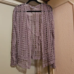 Ljusgrå virkad kofta från South - Säljer en ljusgrå virkad kofta från South i 100% viskos. Koftan har lång ärm, öppen front och knytband framtill. Perfekt att slänga över en topp för en chill och luftig look. Det öppna virkade mönstret ger en bohemisk vibe.