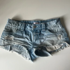Blå jeansshorts från Gina Tricot - Snygga ljusblå jeansshorts från Gina Tricot med slitna detaljer och fransig kant. Klassisk femficksmodell med knapp och dragkedja framtill. Perfekta för varma dagar och en avslappnad look.