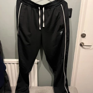 Svarta träningsbyxor från Gymshark L - Svarta träningsbyxor från Gymshark i storlek L. Byxorna har vita ränder längs benen, elastisk midja med vit snörning och Gymshark-logga på låret. Perfekta för gymmet eller chill. Tillverkade i mjukt och lätt material.