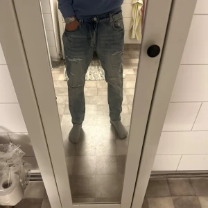 Ljusblå slitna jeans med raka ben - Säljer ett par ljusblå jeans med slitningar och hål på benen. Jeansen har normal passform och raka ben, med klassisk femficksdesign och dragkedja. De är tillverkade i jeansmaterial och har en cool, avslappnad vibe med detaljer som färgstänk och slitna partier. skriv för exakt storlek
