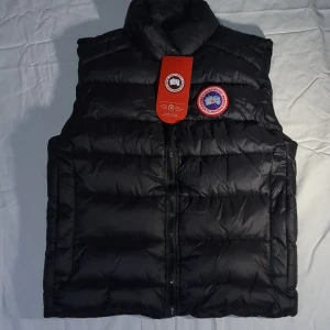 Svart dunväst från Canada Goose - CAN DISKUTERA PRIS!!! Säljer en svart dunväst från Canada Goose med klassisk logga på bröstet. Västen har hög krage, quiltad design och dragkedja framtill. Perfekt för lager-på-lager och håller dig varm under kyliga dagar. Reflekterande detalj i nacken för extra stilpoäng.