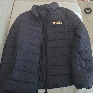 Svart pufferjacka från BOSS - Snygg svart pufferjacka från BOSS med gul logotyp på bröstet och en brun BOSS-detalj på insidan. Jackan har quiltad design, dragkedja framtill och hög krage. Perfekt för kyliga dagar och ger en clean, modern look. 