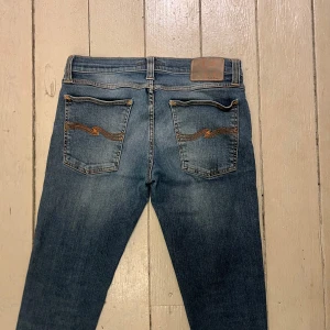 Blå jeans från Nudie Jeans - Snygga blå jeans från Nudie Jeans med klassisk femficksdesign och orange broderi på bakfickorna. Jeansen har en rak passform och är tillverkade i mjukt denimtyg. Perfekta för dig som gillar en tidlös och avslappnad stil.