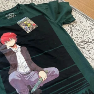 Assassination Classroom anime t-shirt - Grön t-shirt från Assassination Classroom med stor anime-tryck framtill. Motivet visar en karaktär med rött hår och svärd, perfekt för animefans. T-shirten är i bomull och har en klassisk passform med korta ärmar.