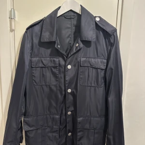 Svart vindjacka från Stenströms - Snygg svart vindjacka från Stenströms med fyra stora fickor framtill, axelklaffar och silvriga knappar. Jackan har klassisk krage och är tillverkad i glansig polyester som ger en clean look. Perfekt för dig som gillar stilren och funktionell design.