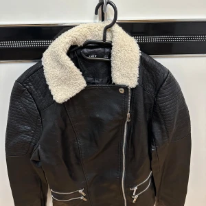 Svart skinnjacka med teddykrage LMTD - Svart skinnjacka från LMTD med vit teddykrage, quiltade detaljer på axlar och ärmar samt fyra dragkedjeförsedda fickor framtill. 