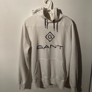 Vit hoodie från GANT  - Vit hoodie från GANT med stor svart logga på bröstet och svarta snören i huvan. Klassisk känguruficka framtill. Perfekt för en chill och stilren look.