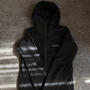 Svart vindjacka Peak Performance - Svart vindjacka från Peak Performance med huva och hel dragkedja framtill. Jackan har diskret logga på bröstet och är tillverkad i ett lätt syntetmaterial som passar perfekt för blåsiga dagar. Snygg, stilren och funktionell design med fickor med dragkedja.