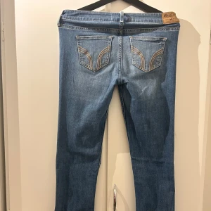 Blå slitna jeans från Hollister - Säljer ett par blå jeans från Hollister med snygga slitningar och hål på knät. Jeansen har klassisk femficksdesign, låg midja och utsvängda ben. Materialet är mjukt denim med lite stretch för skön passform. Bakfickorna har broderade detaljer.