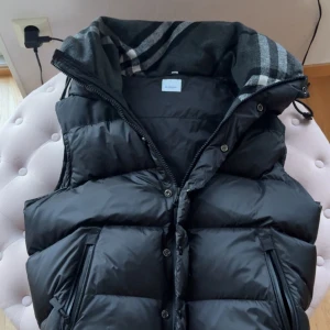 Svart dunväst/jacka från Burberry - Svart dunväst från Burberry med klassisk quiltad design och dragkedja framtill. Västen har en hög krage med rutigt foder och två fickor med dragkedja. På ryggen finns en Burberry London England-logga. Perfekt för lager-på-lager-stil.