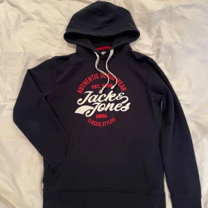 Mörkblå hoodie från Jack & Jones - Snygg mörkblå hoodie från Jack & Jones med vit och röd tryckt logga på bröstet. Tröjan har huva med vita snören och en stor magficka. Perfekt för chill dagar och streetstyle.