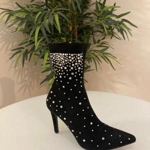 Svarta stövletter med strass och klack - Svarta stövletter i mockaliknande material med spetsig tå och hög klack. Skorna är dekorerade med glittrande strass-stenar i olika storlekar som ger en riktigt lyxig och trendig vibe. Perfekta för dig som vill sticka ut och addera lite extra bling till outfiten.