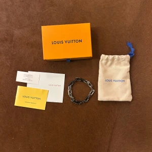 Louis Vuitton silverfärgat armband - Snyggt armband från Louis Vuitton i silver- och svartfärgad metall med kedjelänkar och ingraverade LV-loggor. Kommer med original ask, dustbag och äkthetskort. Perfekt accessoar för dig som vill sticka ut med exklusiv stil.