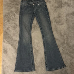 True Religion bootcut jeans blå - Snygga blå jeans från True Religion med klassiska kontrastsömmar och bootcut-modell. Jeansen har låg midja och ikoniska bakfickor med broderad logga. Tillverkade i slitstarkt denim med lätt tvättad look och markerade benslut. På tredje bilden syns det att de är en aning slitna men annars som nya. 