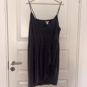 Svart omlottklänning med volang - Svart kortklänning från H&M med tunna axelband och v-ringning. Klänningen har omlottdetalj framtill och volang längs nederdelen. Tillverkad i mjuk polyester med en lätt glansig finish. Perfekt för dig som gillar stilrena och enkla plagg med en twist.