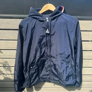 Mörkblå vindjacka från Moncler - Snygg mörkblå vindjacka från Moncler med huva och dragkedja framtill. Jackan har diskreta fickor med tryckknappar och Monclers logga på dragkedjan. Perfekt för blåsiga dagar och enkel att matcha med det mesta.