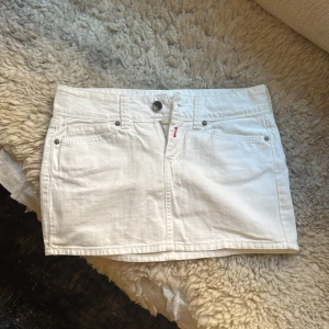 Vit jeanskjol från Replay, storlek 27 - Vit kort jeanskjol från Replay med klassisk femficksdesign, broderad logga bak och snygga dragkedjor på bakfickorna. Kjolen har knapp- och dragkedjestängning framtill och är tillverkad i bomull med en touch av stretch för extra komfort.