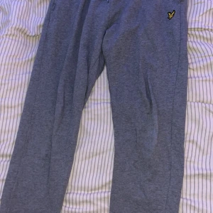 Grå mjukisbyxor från Lyle & Scott - Grå mjukisbyxor från Lyle & Scott med klassisk gul fågel-logga broderad på vänster sida. Byxorna har dragsko i midjan och är tillverkade i mjukt bomullsmaterial. Perfekta för chill eller träning, med normal passform och fickor på sidorna.