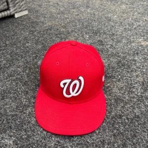 Röd New Era Washington Nationals keps - Snygg röd keps från New Era, modell 59FIFTY, med Washington Nationals-logga broderad i vitt och svart framtill. Kepsen har en rak skärm och broderade ventilationshål. Tillverkad i 100% polyester och har MLB-logga baktill.
