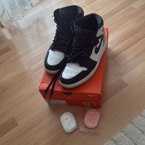 Nike Air Jordan 1 svart/vit 42.5 - Nike Air Jordan 1 i svart och vitt med klassisk hög siluett. Skorna har svart swoosh, perforerad tåbox och kommer med extra skosnören i vitt och rosa. Tillverkade i skinn och syntetmaterial, med snörning och rund tå. Perfekta för dig som gillar streetwear och sneakerskultur.