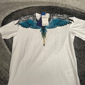 Vit t-shirt Marcelo Burlon med vingar - Snygg vit t-shirt från Marcelo Burlon County of Milan med ikoniskt vingtryck i blått, svart och grönt över axlar och bröst. Klassisk passform och rund hals. Perfekt statement-plagg för dig som vill sticka ut.