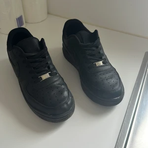 Nike Air Force 1 helsvarta sneakers - Säljer ett par klassiska Nike Air Force 1 i helsvart skinn. Modellen har lågt skaft, perforerad tå och svarta snören med silverfärgad detalj. Swoosh-loggan på sidan och diskret Nike Air-märke på plösen. Perfekta för dig som gillar en clean och tidlös look.