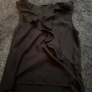 Svart volangblus från Zara Basic XS - Säljer en svart ärmlös blus från Zara Basic Collection i storlek XS. Toppen har en snygg volangdetalj längs halsringningen och framtill, vilket ger en trendig och feminin look. Tillverkad i ett lätt och mjukt tyg som känns skönt mot huden.