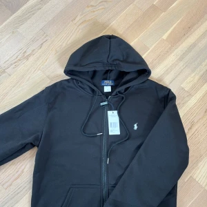 Svart hoodie från Polo Ralph Lauren - Svart hoodie från Polo Ralph Lauren med dragkedja framtill och klassisk huva med snörning. Tröjan har det ikoniska broderade logotypen på bröstet och är tillverkad i mjuk bomullsmix. Perfekt för en chill och stilren look.