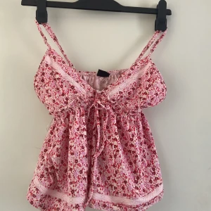 Blommig rosa linnetopp från Gina Tricot - Supersöt rosa linnetopp från Gina Tricot med blommigt mönster i rött och ljusrosa. Toppen har smala axelband, spetsdetaljer och knyt framtill. Perfekt för varma dagar och ger en romantisk vibe till din outfit.
