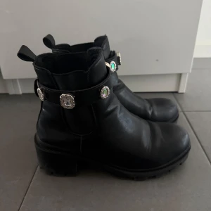 Svarta boots med smyckade spännen - Snygga svarta boots i skinn med chunky klack och rund tå. Har tappat en diamant grej på vänstra men det syns knappt! Storlek 39