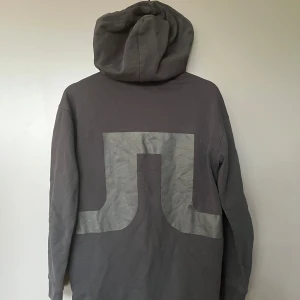 Svart hoodie från J.Lindeberg XS - Svart hoodie från J.Lindeberg i storlek XS. Tröjan har en stor grå logga på ryggen, huva med snörning och klassisk känguruficka framtill. Perfekt för en chill och stilren look. Mjuk och skön bomullskvalitet.