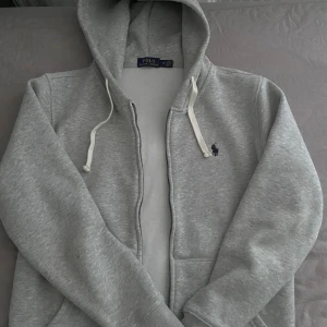Grå hoodie från Polo Ralph Lauren M - Grå hoodie från Polo Ralph Lauren med klassisk broderad logga på bröstet. Tröjan har huva med vita snören, dragkedja framtill och två fickor. Mjuk och skön bomullsblandning, perfekt för chill dagar eller när du vill ha en clean look.