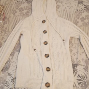 Vit stickad kofta med huva No Name - Mysig vit stickad kofta från No Name med stora bruna knappar och huva. Koftan har kabelstickat mönster framtill, ribbade muddar och dragsko i huvan. Perfekt för kyliga dagar och enkel att matcha med allt.